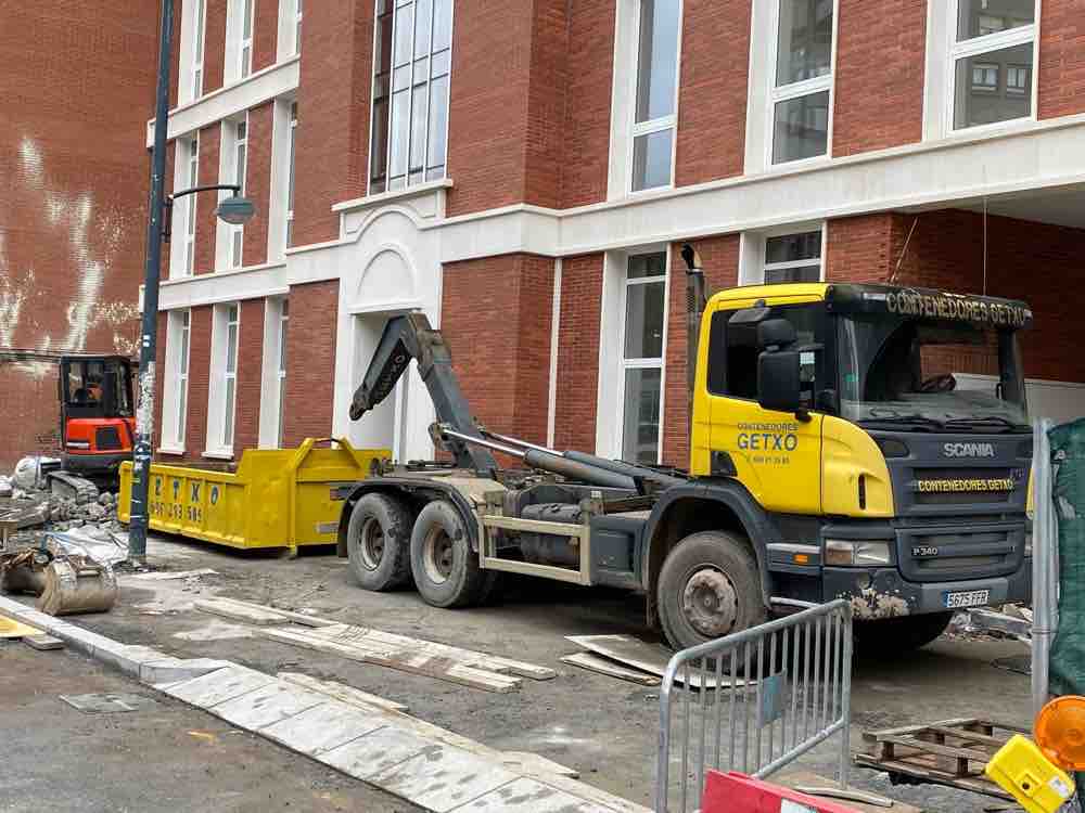 Camión Scania amarillo de Contenedores Getxo con brazo de gancho junto a un contenedor de escombro, frente a un edificio de ladrillo en obras con una miniexcavadora